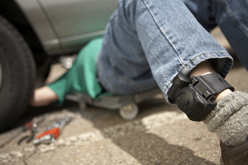 A man works under a car, wearing a BI TAD Transdermal Alcohol Detector on his ankle to detect alcohol consumption noninvasively.