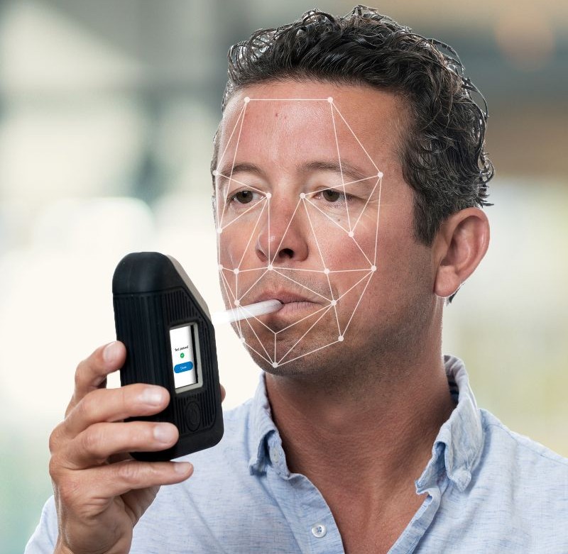 Man using BI SoberTech alcohol monitoring device, blowing into straw with facial biometrics overlay verifying client identity during alcohol test.