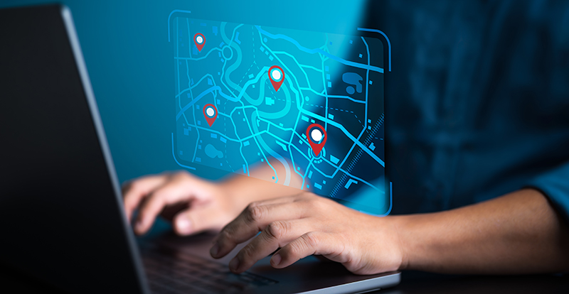 A corrections officer working at a laptop computer, viewing a digital map with location pins and tracking data overlays. The image illustrates modern location tracking systems in community supervision.
